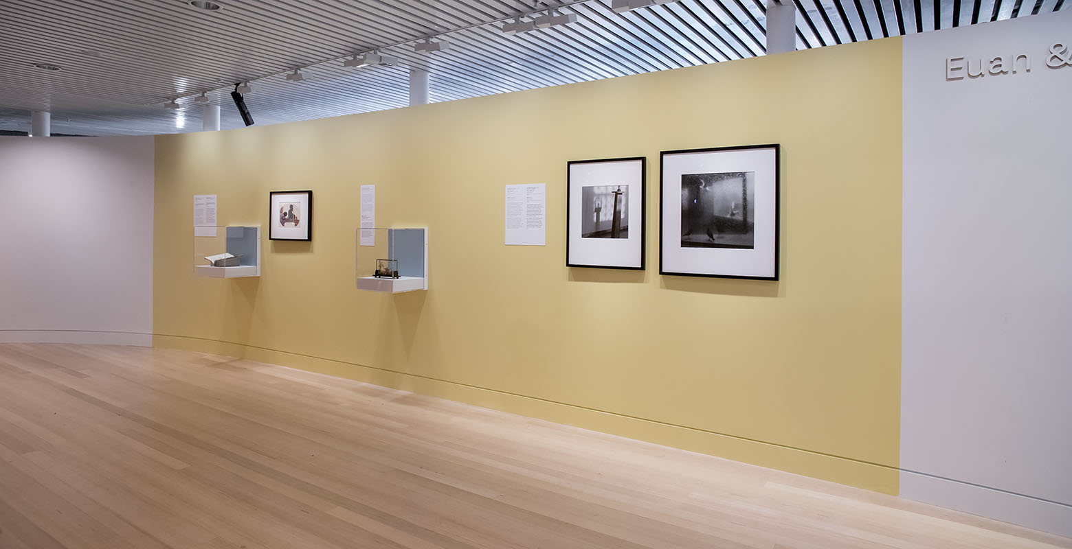 Two photographs are hung on a yellow wall, with a book and a wooden Chinese garden scene housed in display cases