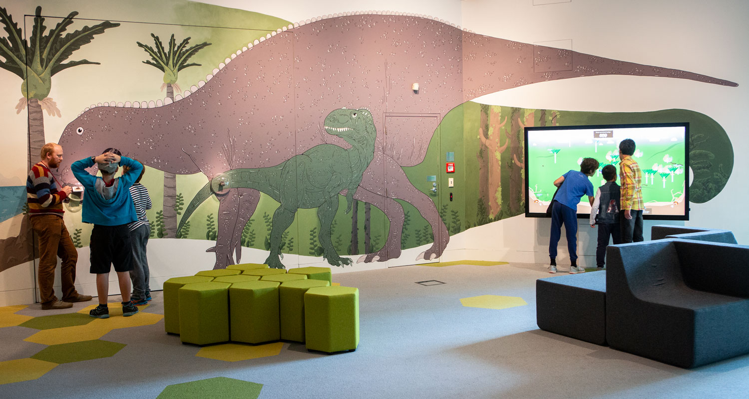 People in a room with large dinosaurs painted on the walls