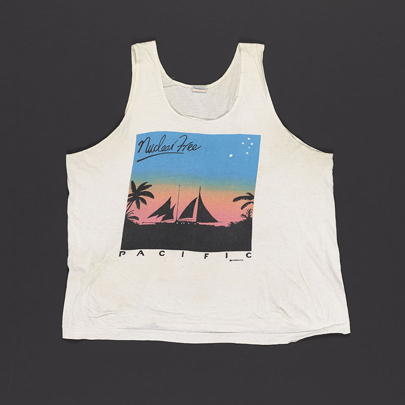 White singlet with a vibrant graphic on it of a silhouetted boat on the ocean surrounded by trees, a pink sky blending into a blue sky, and the words Nuclear free Pacific written on it