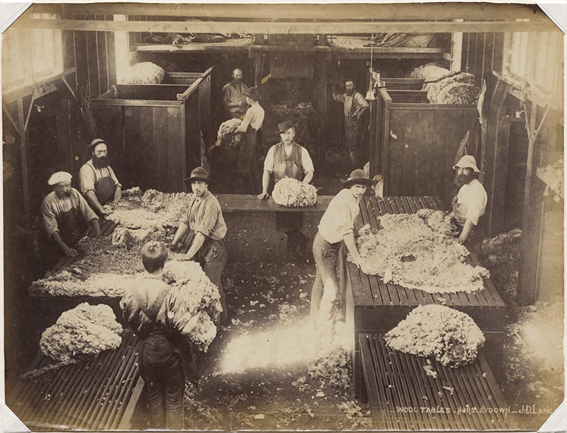A sepia photo of a woolshed with ten people surrounding big tables with shorn wool sitting on them