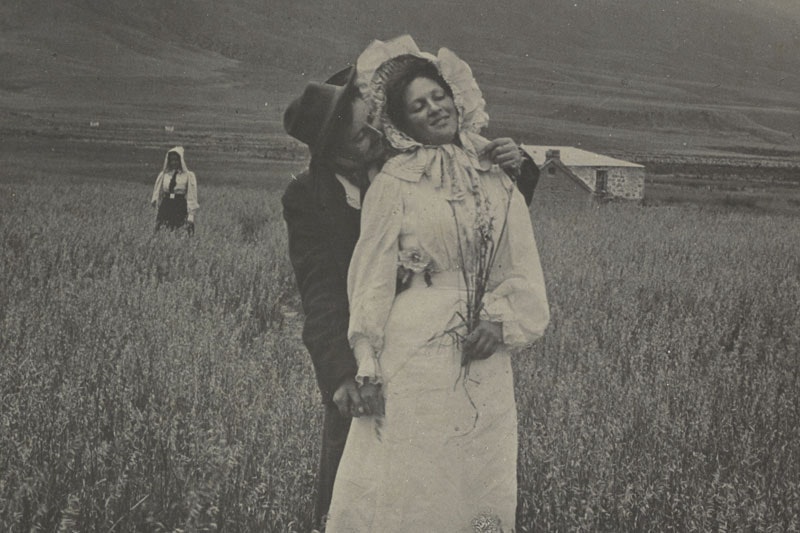 A man hugs a woman from behind in a field. She holds a bunch of flowers. Behind them a woman stands in isolation. Further back, there’s a stone house and hills