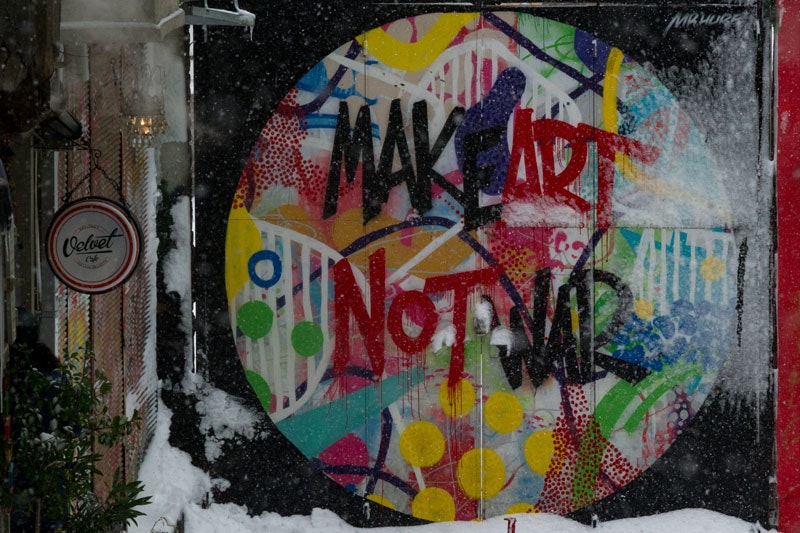 Photo of a wall with graffiti on it. The graffiti is a colourful circle and inside it is written "Make Art Not War". It has been snowing and snow has settled on the ground at the wall
