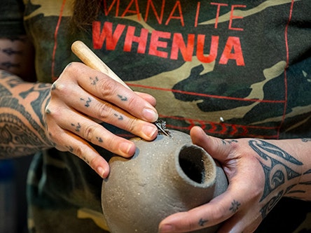 Stevei Houkamau in her studio, December 2020. Photo by Daniel Crichton-Rouse. Te Papa (170298) A woman's tattooed hands are holding a clay vase – she is carving shapes into it.