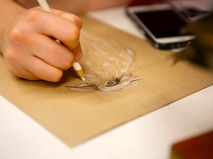 Te Taiao Nature After Dark 2019. Photo by Jo Moore. Te Papa (138587) A close up of a hand drawing a bird on a piece of brown paper.