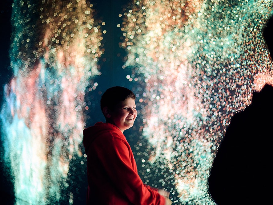 Distortions in Spacetime. Marshmallow Laser Feast: Works of Nature, 2023, ACMI. Photo © Eugene Hyland A child is smiling up at someone with them. They are both in front of a gigantic screen with dots of light and swirling patterns.