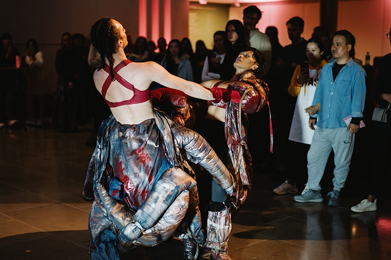 Body Island performing at Auckland Art Gallery, October 2025. Photo by David St George. Courtesy of Auckland Art Gallery Toi o Tāmaki Two performers wearing costumes like puffy exoskeletons hold hands as they lean back. Viewers stand around them, watching on.
