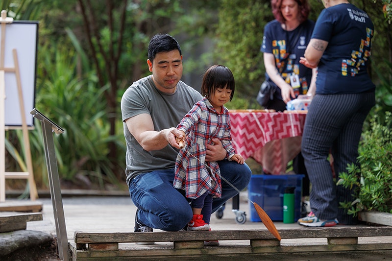 Raumati Whānau Day 2025. Photo by Jo Moore. Te Papa (257122) Man with little girl playing and whirling a wooden instrument outside.