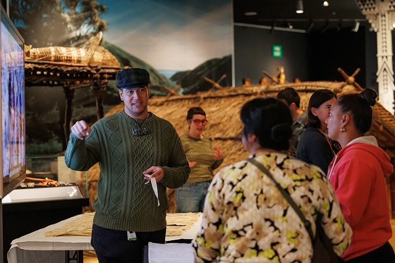 Mātauranga Māori Curator Isaac Te Awa. After Dark: Mātahi o Rongo 2025, photo by Jo Moore. Te Papa (261982) A male Curator is standing in an exhibition facing the camera, wearing a flat cap, green sweatshirt and a hei tiki pounamu taonga around his neck. He speaks to two women who have their back to the camera, holding a small piece of material in his hand.