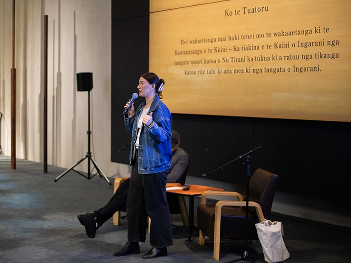 After Dark: Ka tangi te Pīpīwharauroa 2025, Photo by Renati Waaka Young woman speaking with a microphone in the Signs of a nation exhibition at Te Papa.