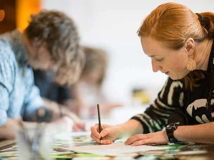 Zine-making workshop with Kerry Ann Lee. Photo by Michael Hall. Te Papa (32002) People are working with pencils and paper on a table. There is one woman in focus at the front and other people blurred in the background.