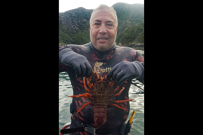 A man in a wetsuit is holding a crayfish up in front of himself. He is looking at the camera.