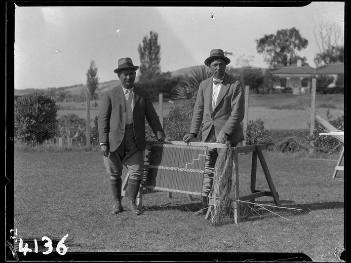 Sir Apirana Ngata (left) and Sir Peter Buck (right) on an ethnology expedition. Photo by James McDonald, 1923. Te Papa (MU000523/006/0005) Black and white photo of two people standing in a field next to a piece of weaving