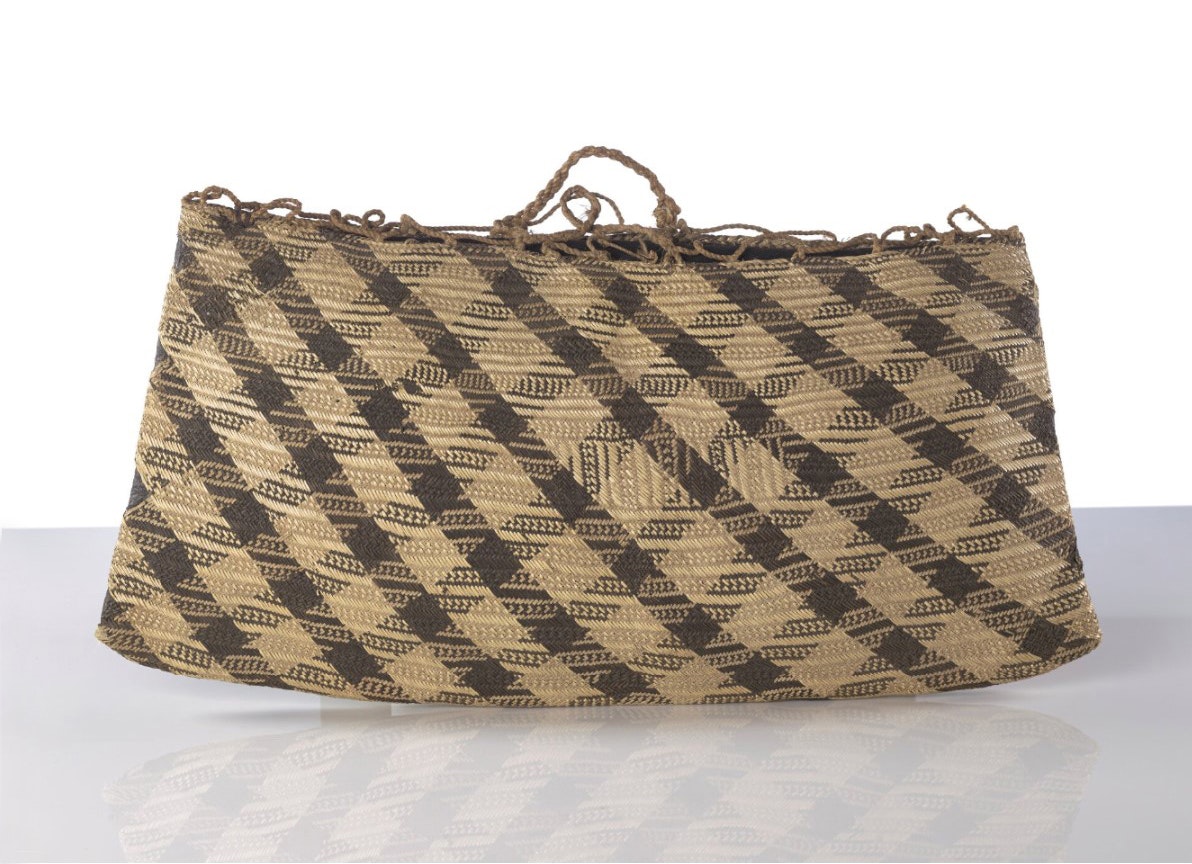 A flax bag with two handles and dark brown patterns woven into it. On a glass reflective surface and a white background.