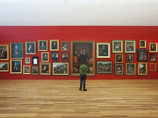 The portrait wall in Tūrangawaewae: Art and New Zealand, 2018. Photograph by Michael O’Neill. Te Papa A man stands looking at a red wall featuring paintings hung in the salon style