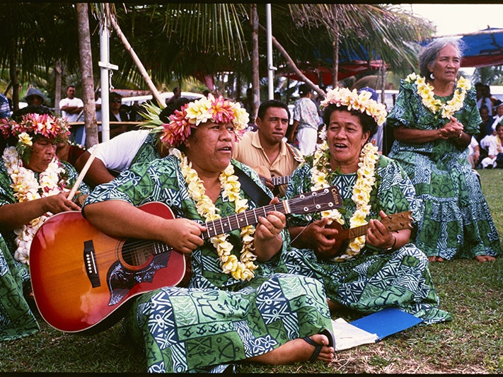 ma_i435229_jowitt_tile.jpg Women wearing the same dresses and headwear are sitting in a group. One is playing the guitar and one is singing.
