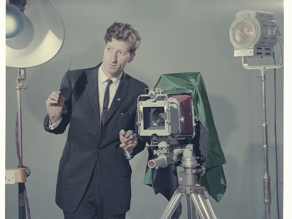 studio_photography_ma_i031167.3000x3000.jpg A man in a suit stands between stage lights and has an old-fashioned camera on a tripod next to him