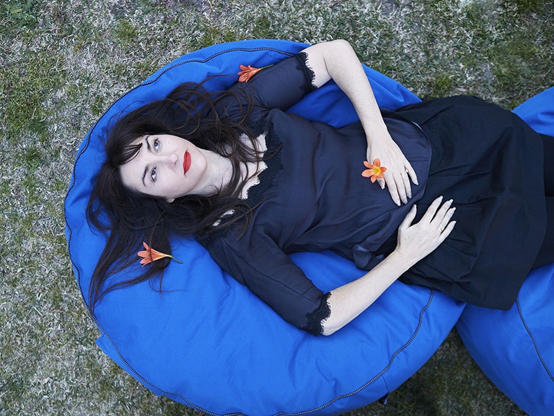 Nike Savvas, 2017. Photo by and courtesy of Zan Wimberley Nike Savvas, dressed in a dark blue dress, lies on a lighter blue beanbag on the grass, holding and surrounded by orange flowers