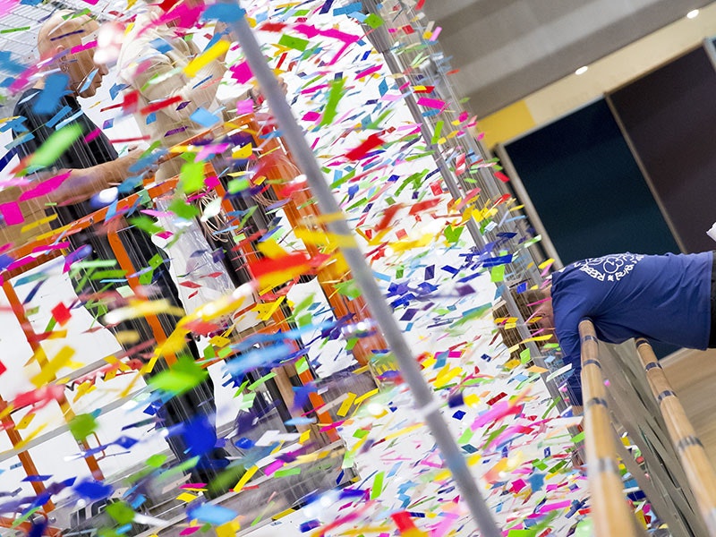 Installing Nike Savvas, Finale: Bouquet, 2019. Photo by Maarten Holl. Te Papa A person on a scissor lift is obscured by a dense collection of strands of colourful pieces of plastic hanging from the ceiling while another person looks over a bridge beside the work down towards the floor, where the work continues