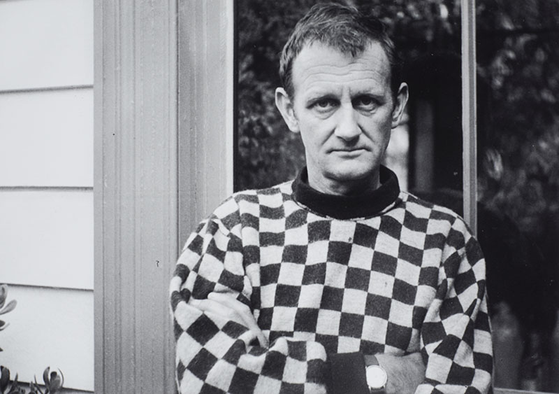 Colin McCahon stands outside a house in front of a large window, wearing a checkered shirt, with his arms folded
