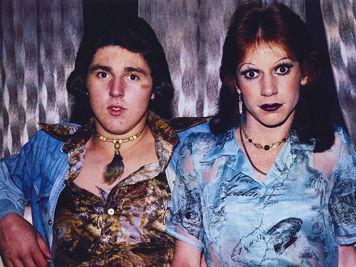 Donna Terri and friend at Mojos. Auckland, by Fiona Clark, 1975. Te Papa (O.043785) Two people seated look into the camera – one a man in an open shirt with a forest on it and denim jacket, the other a man in makeup wearing a blue shirt with a map of New Zealand on it