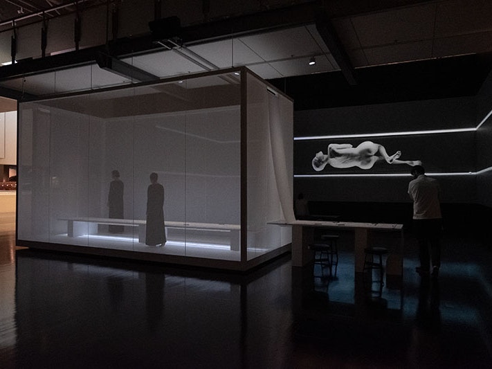 Installation view of Lemi Ponifasio, MAU: House of Night and Day, 2019. Photo by Rachael Hockridge. Te Papa A man walks around a space that contains a box with two women inside and a massive projection on the wall of a woman contorting her body