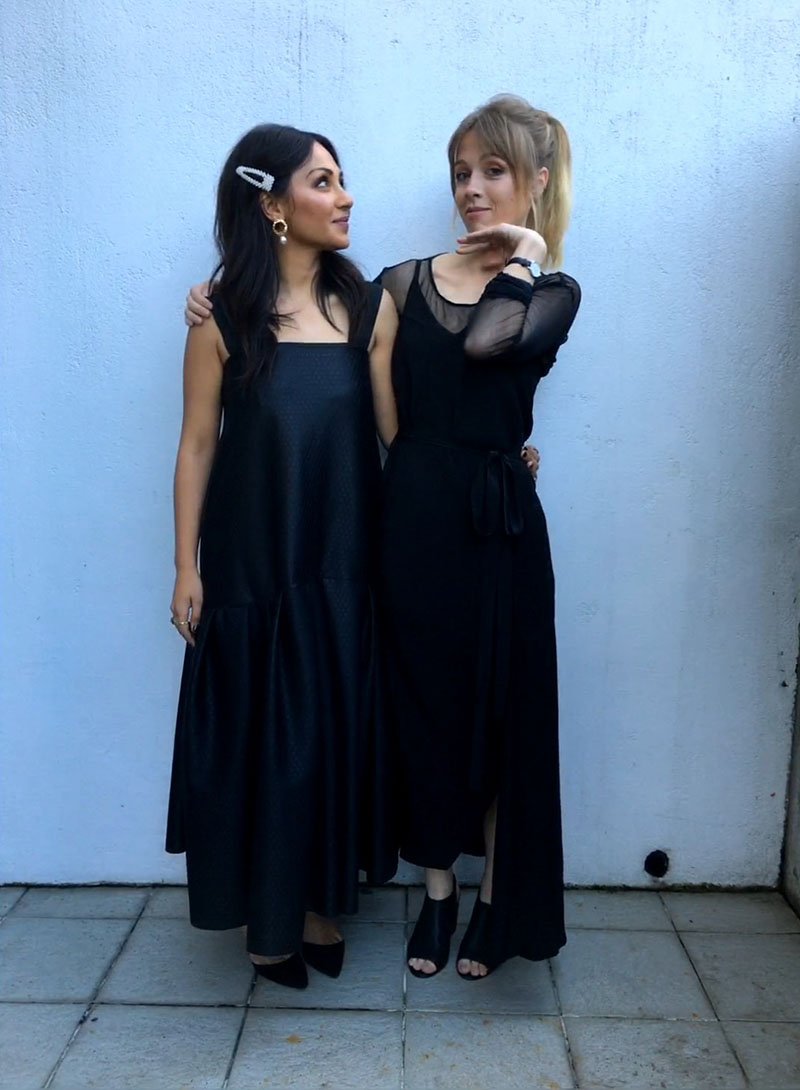 Two women in elegant black evening dresses pose for a photo against a concrete wall