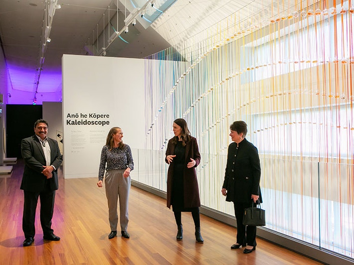 Kaihautū | Māori Co-leader Arapata Hakiwai, Tumu Whakarae | Chief Executive Courtney Johnston, and Chair of the Te Papa Board Dame Fran Wilde with Prime Minister Jacinda Ardern in Te Papa’s Toi Art ahead of the PM’s funding announcement. Photo by Jo Moore Four people stand talking in an art gallery with a large rainbow of colours behind them