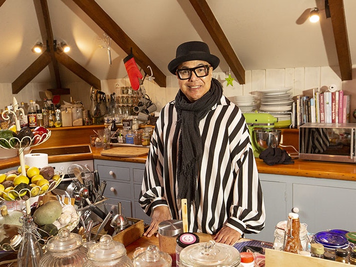 Uncle Pare in his kitchen, 2020. Photo by Norm Heke. Te Papa Uncle Pare stands in his kitchen. He wear a black and white striped shirt, black scarf, glasses, and a black brimmed hat. His kitchen is overflowing with a well organised array of bottles, utensils, plates, and jars