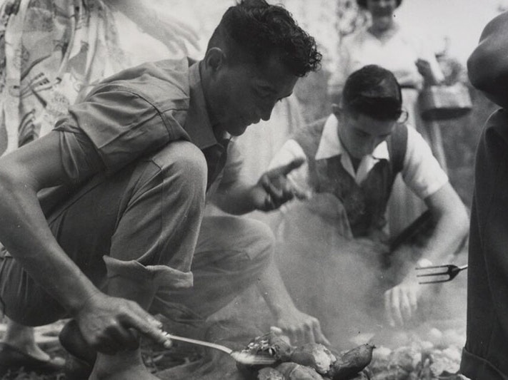 Hāngī, Hokianga, 1957, by Eric Lee-Johnson, Hokianga Harbour. Purchased 1997 with New Zealand Lottery Grants Board funds. Te Papa (O.006431) Two young men work to prepare food for a hāngī. The first is using a spatula to check on food from the hāngī while the second is lifting the food from the pit. Three women and another man are in sight around the hāngī pit.
