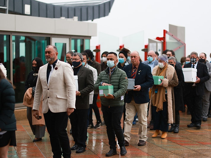 Maui Solomon and Te Arikirangi Mamaku lead the delegation on to Te Papa’s marae, carrying the karāpuna (Moriori anscestors). Repatriation Ceremony, July 2022. Photo by Te Papa (206620) A man in a white jacket leads several people into a building. Many of them are carrying green boxes with human remains in them