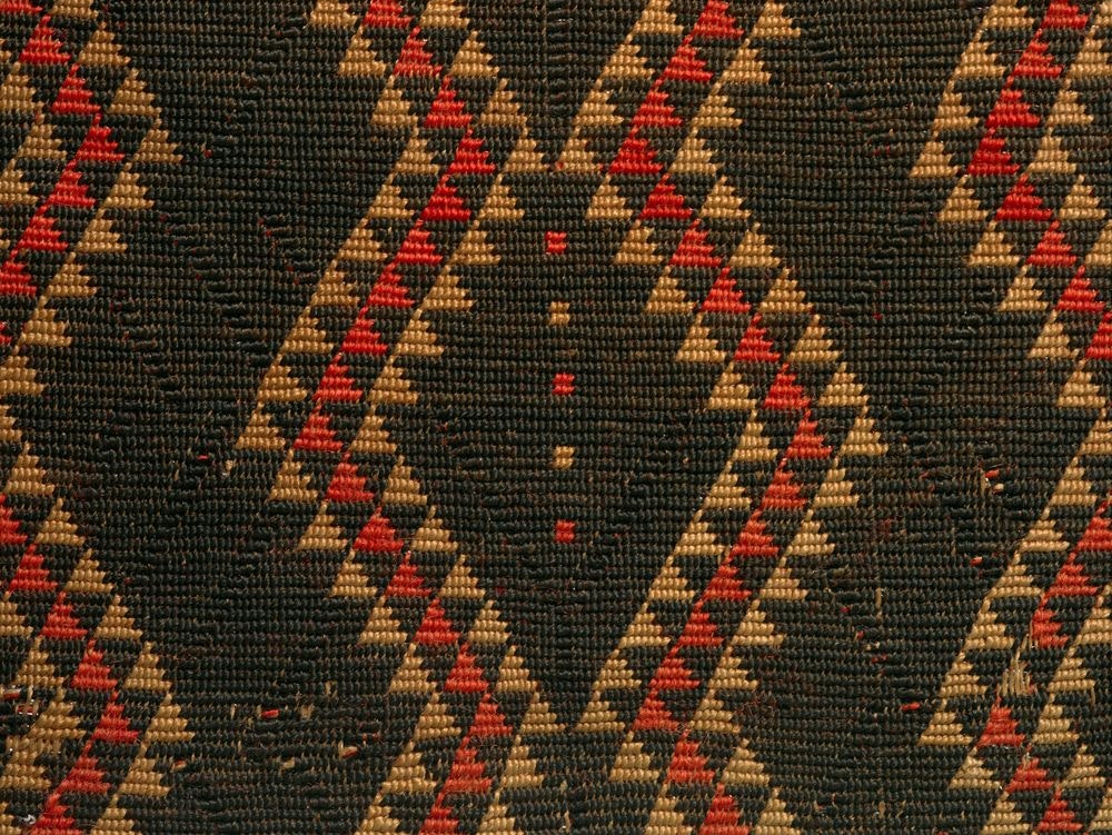 Close up of a cloak, showing the detail of its design. It is predominantly black with red and earth coloured triangles forming a wave across the cloak