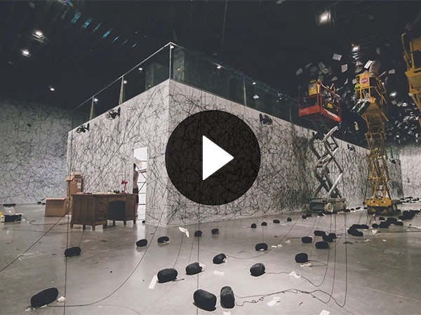 Still from TIMELAPSE CHIARU SHIOTA, 28 Oct 2014, by Wally Sanz. Still from a video. It shows a large pavilion in the middle of a room, three scissor lifts, and multiple strands of black wool hanging from the ceiling mid-installation
