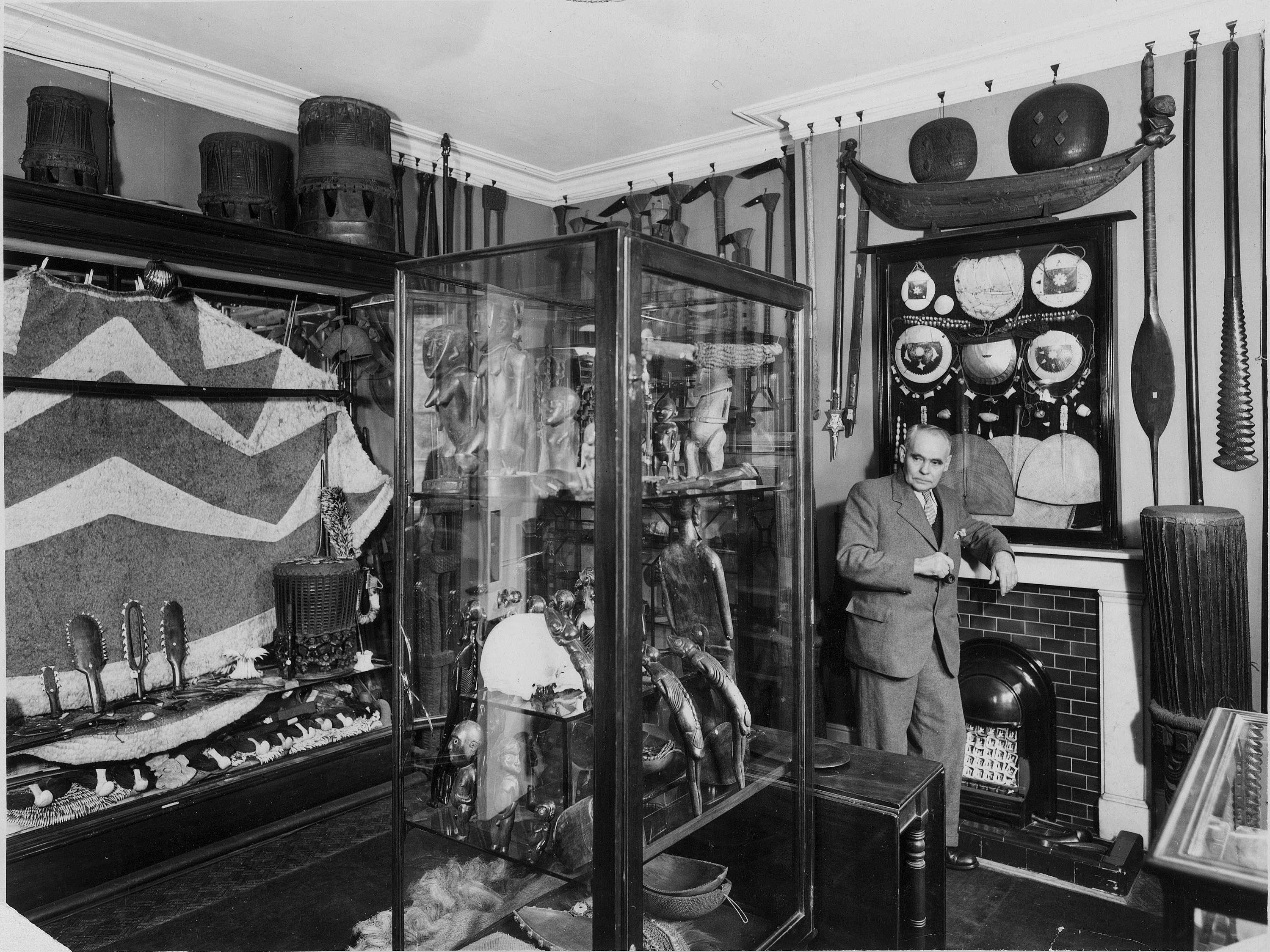 W.O. Oldman with his collection, at the Clapham villa, circa 1940, black and white photograph, gelatin silver print, Hobbs, Offen & Co Ltd. Te Papa (O.027324) A black and white photo with an older man standing in the middle of a lot of traditional Pacific art pieces