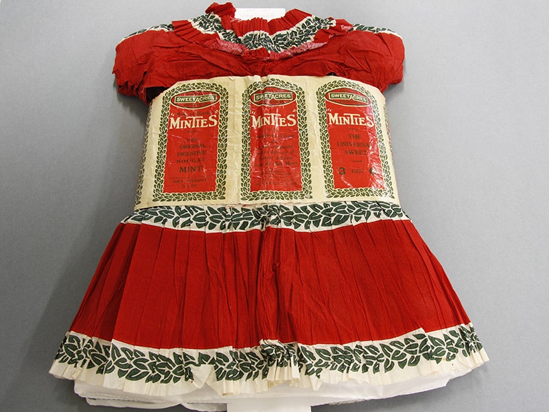 Costume, child's (dress and hat made out of Minties labels), circa 1927, maker unknown. Gift of Shelley Venimore (née O'Brien), 2011. CC BY-NC-ND 4.0. Te Papa (GH017183) A red child's dress with Minties lollies marketing on it. It's laid flat on a grey background.