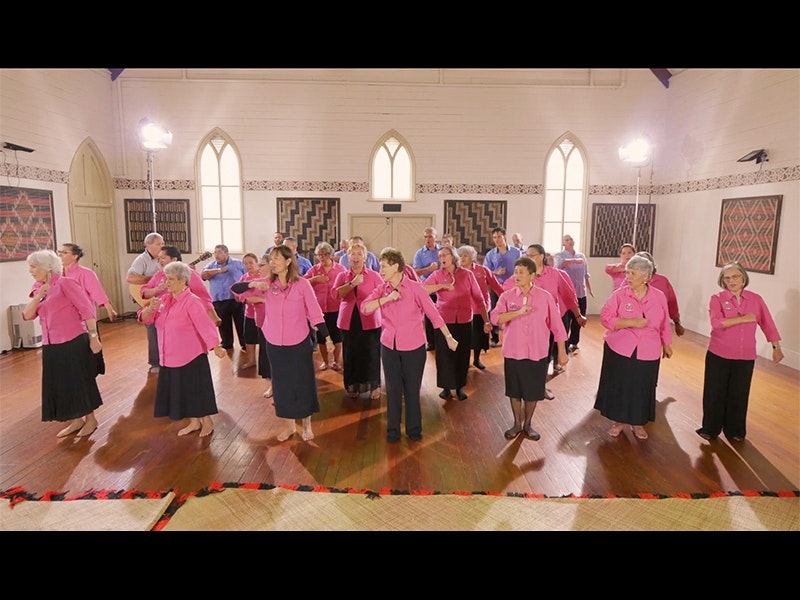 Photo courtesy of Auckland Anglican Māori Club A group of older people wearing pink shirts and blue shirts dancing in formation inside a church hall