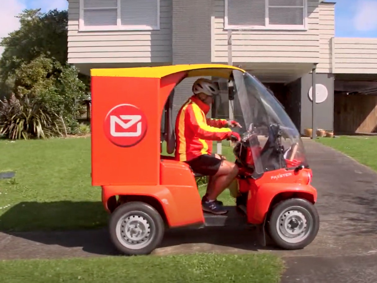 using_electric_vehicles_-_nz_post_nationwide.jpg A person driving a red and yellow electric vehicle in front of a house
