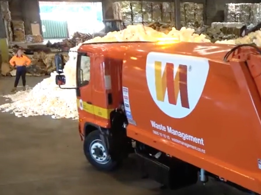 Video still from Waste Management NZ, nationwide. Te Papa A waste management truck is in a shed with mounds of empty plastic milk bottles and a person standing in the background