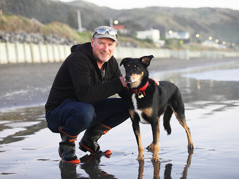 Mike White, pictured with Cooper. Photo by Nikki Macdonald A man crouching on a beach with his hand on a dog