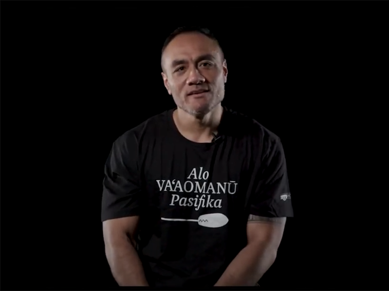 Still from Sadat Muaiava talking about tatau. Te Papa A man in a black t-shirt with Samoan writing on it sitting in a black room