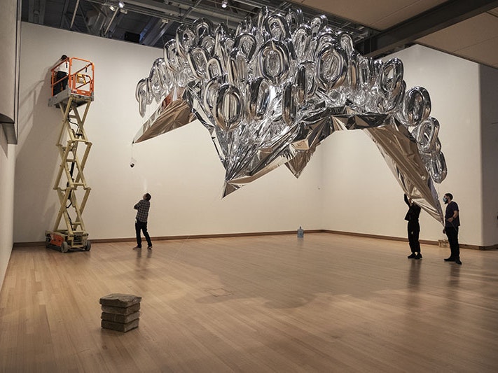 Installation of Mikala Dwyer's The silverings, 2020. Photo by Daniel Crichton-Rouse, Te Papa Three people on the floor and one person in a scissor lift install a large bunch of silver helium balloons in a large room