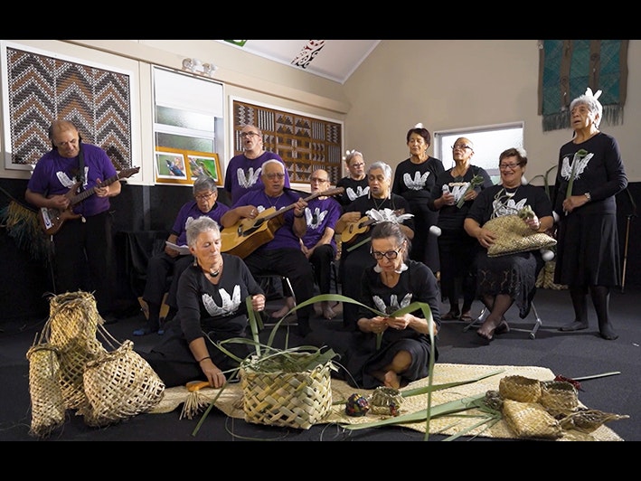 Still from Taikura Kapa Haka 2022 video series Two women sitting in the foreground weaving flax with several people standing behind her singing a song. Some of those are playing a guitar and a ukulele