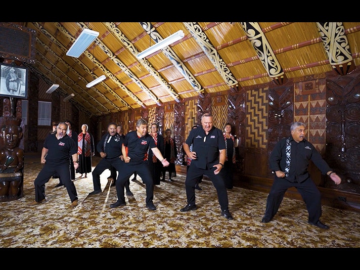 Video still from Taikura Kapa Haka 2022. Te Papa Six men are performing a cermonial dance in a carved meeting hall. There are women singing at the back