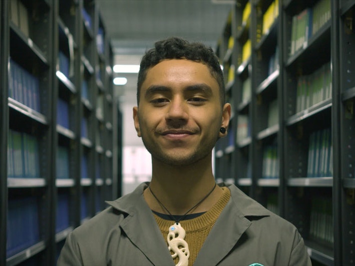 Still from Ngā Taonga Tuku Iho #3: Tukuna Kia Rere, 2022, by Pikihuia Haenga and Elise Lanigan. Image courtesy of Kahu Kutia A man stands looking directly into the camera in the Ngā Taonga archives, flanked by shelving