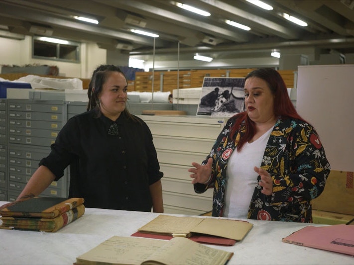 Still from Ngā Taonga Tuku Iho #4: Repatriation, 2022, by Pikihuia Haenga and Elise Lanigan. Image courtesy of Kahu Kutia Two women stand talking in a museum storeroom, surrounded by storage cabinets