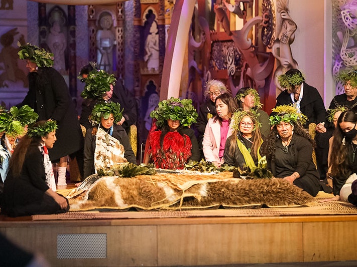 The tūpuna and kārapuna (ancestors) laid on the atamira (stage), 2 Oct 2022. Te Papa A group of woman sit on a stage surrounding ancestral remains, which are covered by feathered cloaks