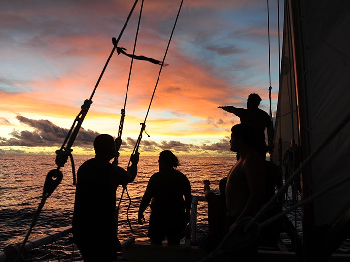 Crew sailing back to Aotearoa from Rarotonga on Ngahiraka-mai-Tawhiti during the Waka Tapu voyage (2013), photo courtesy of Danee Hazama A sunset photo with four crew on a boat in silhouette against the sea and setting sun.