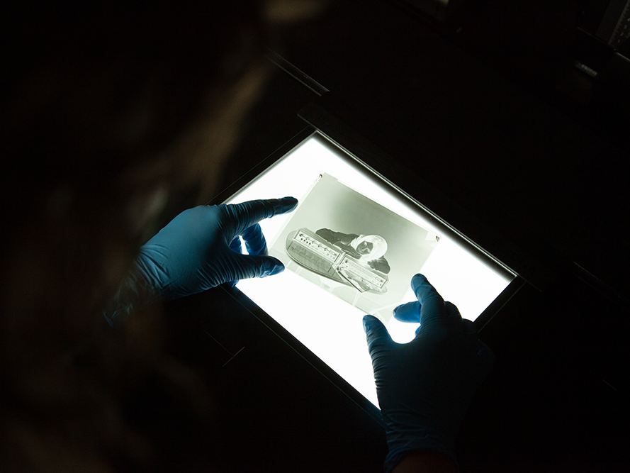 Inspecting a negative on a lightbox. Photo by Melissa Irving, Te Papa A dark photo of two hands wearing blue gloves holding a photographic negative on a lightbox. The negative shows a woman leaning on a piece of machinery