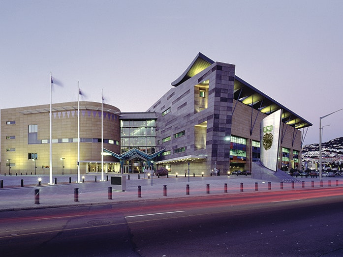 Te Papa at dusk, 2015. Photo by Te Papa Imaging. Te Papa (108057) A large building that has oddly shaped walls photographed at dusk. There are three flags outside the entrance.