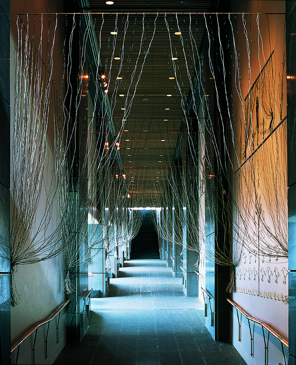 A long passageway with intermittent tall windows and a staircase at the end. There are silvery strings strung and pinned back like curtains along the passage.