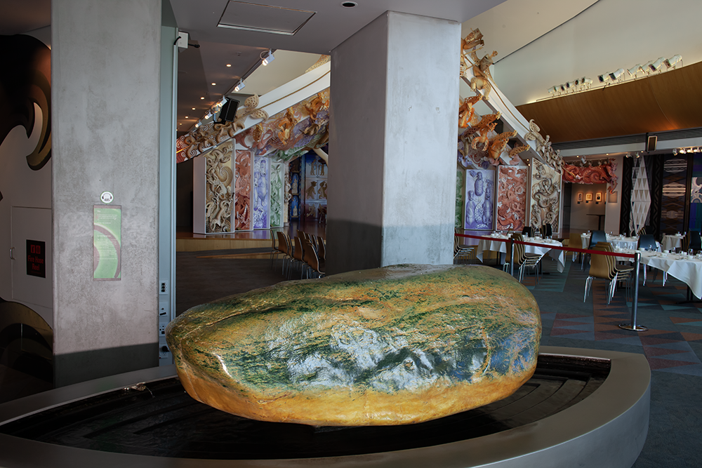 A large boulder in a room. It is partly green and partly yellow. It's sitting in a large wet bowl with running water.